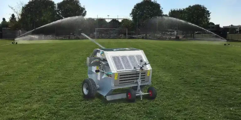 Un irrigatore semovente costituito da una scatola in metallo con ruote, dotato di pannello fotovoltaico. Location campo da calcio.